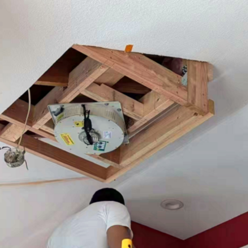 Man installing a ceiling fan within a wooden frame during home renovation, showcasing the structure and wiring.